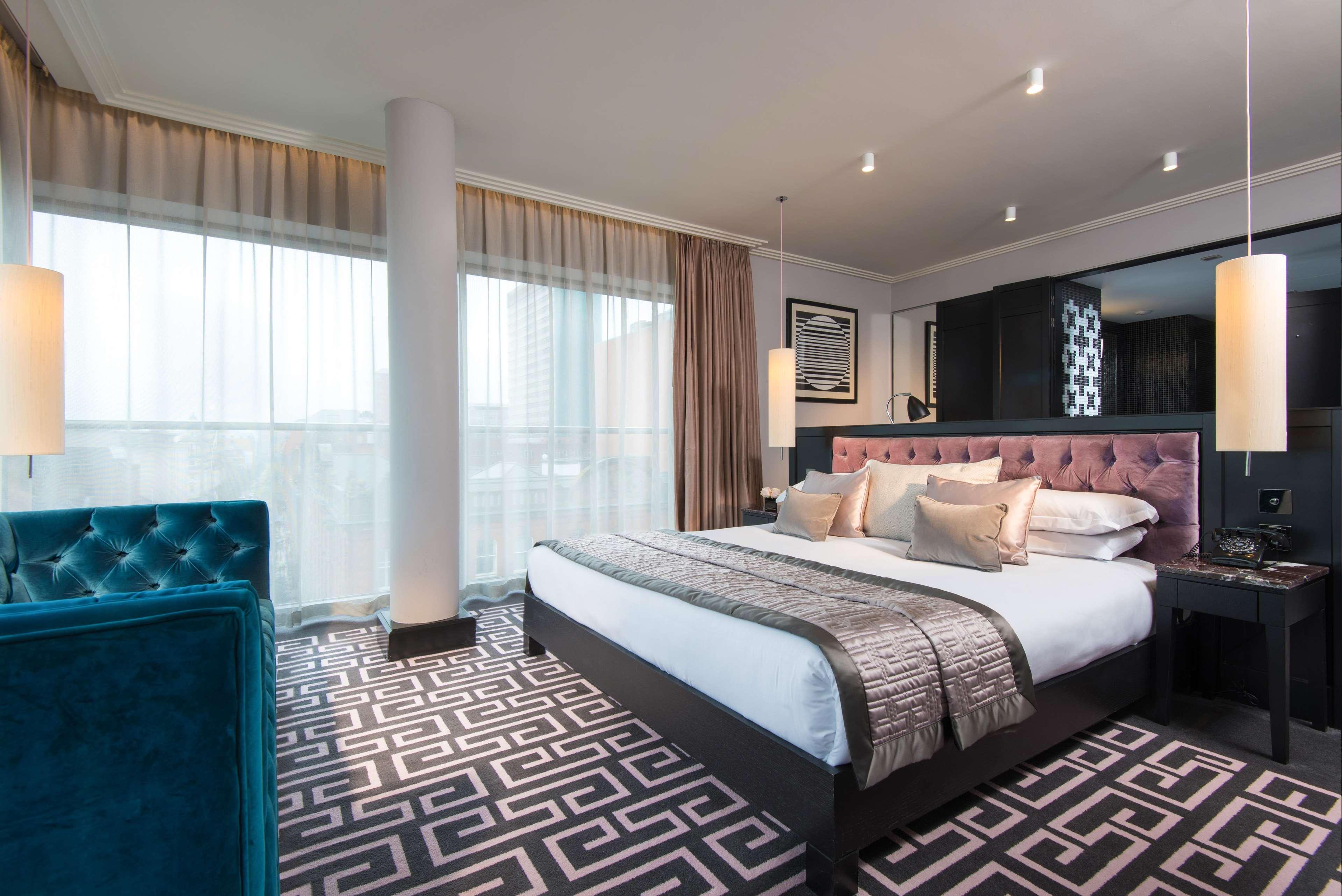 The Fitzwilliam Hotel Belfast Luaran gambar The photo showcases a modern hotel room. It features a large bed with plush pillows and a stylish headboard. The room has large windows with sheer curtains, allowing natural light to filter in. The floor is covered with a patterned carpet that contra